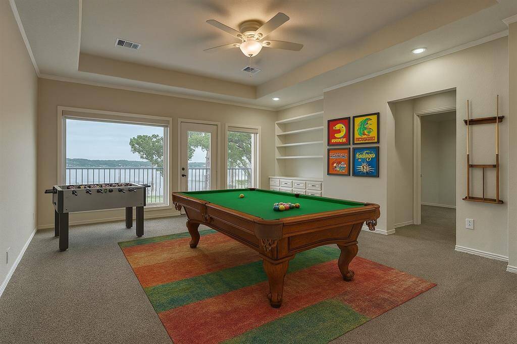 The game room features a wall of windows overlooking the lake and back yard. Built-in cabinetry offers convenient storage for games, media, or a TV. This versatile space can serve as a second living room, game room, exercise area, or playroom. Just beyond, you'll find an additional guest bedroom with an en-suite bath for added comfort and privacy. Virtually staged to show furniture placement.