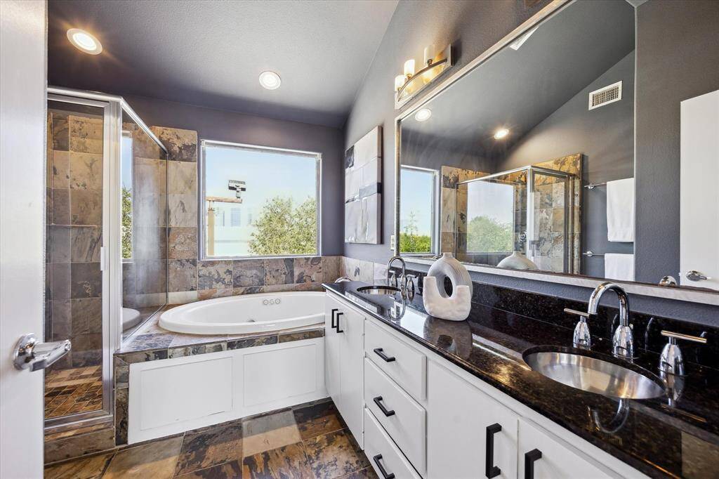 The stunning ensuite bath invites relaxation with a luxurious jetted tub, separate walk-in shower, and extended dual-sink vanity with granite counters, a full framed mirror and chic vanity lights above. Slate tile flooring and surround add natural texture and warmth, transforming the space into a serene, spa-like retreat.