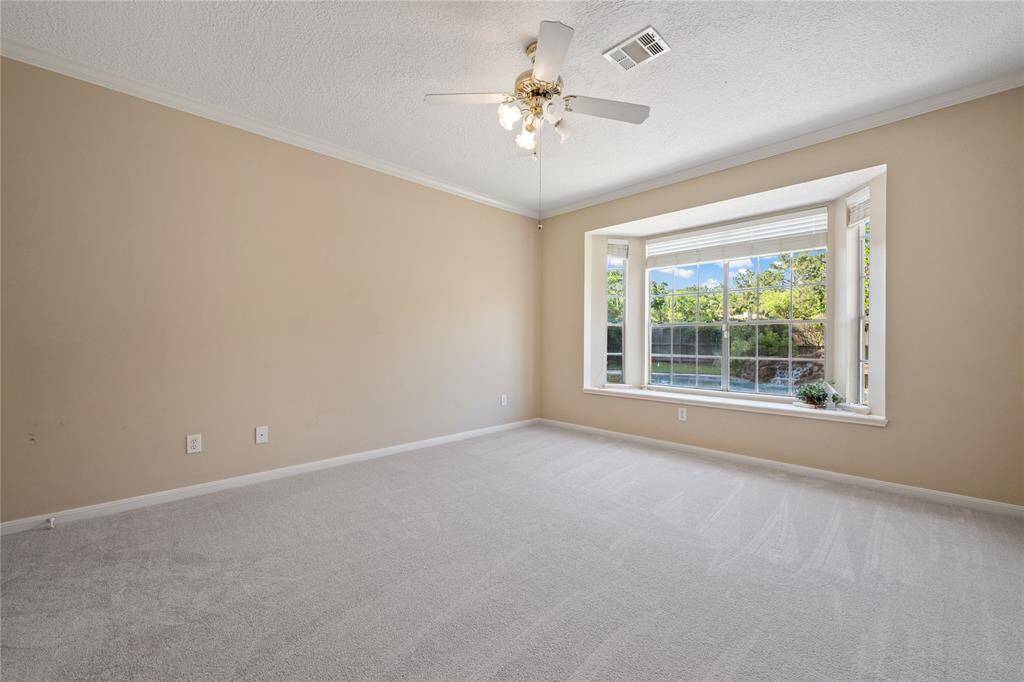Bright and spacious primary bedroom with neutral walls, carpeted flooring, a ceiling fan, and a large bay window offering plenty of natural light and a view of greenery outside.