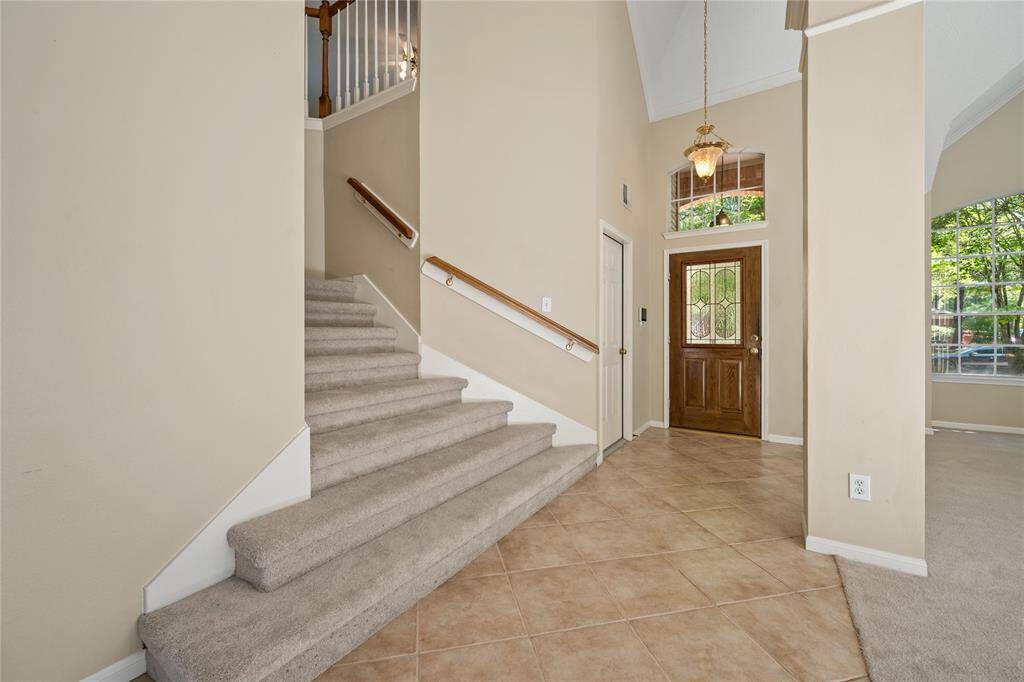 This photo showcases a bright entryway with high ceilings, a wooden front door with glass accents, and a carpeted staircase. The space features neutral tile flooring and large windows, creating an inviting atmosphere.