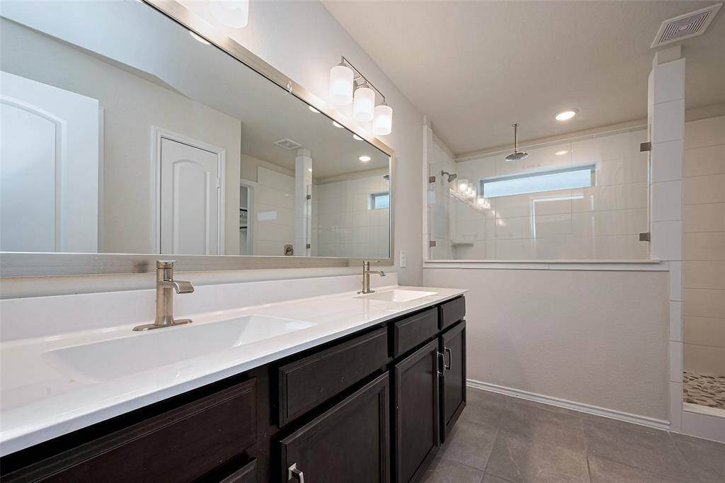 Finished with sleek tile, the bathroom includes a separate shower, dual-sink vanity with extended countertop, wide mirror, and chic lighting—balancing form and function beautifully.