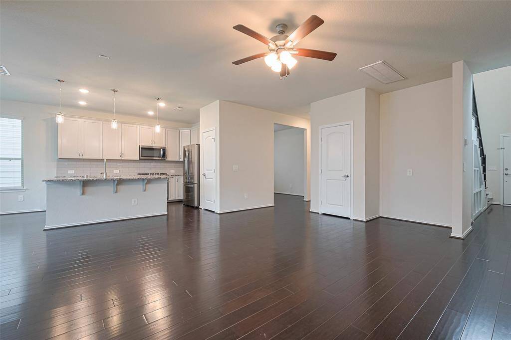The living area feels expansive and open, enhanced by multiple windows, a ceiling fan, and the rich contrast of deep-toned flooring against soft, neutral walls—seamlessly connected to the kitchen for effortless flow.