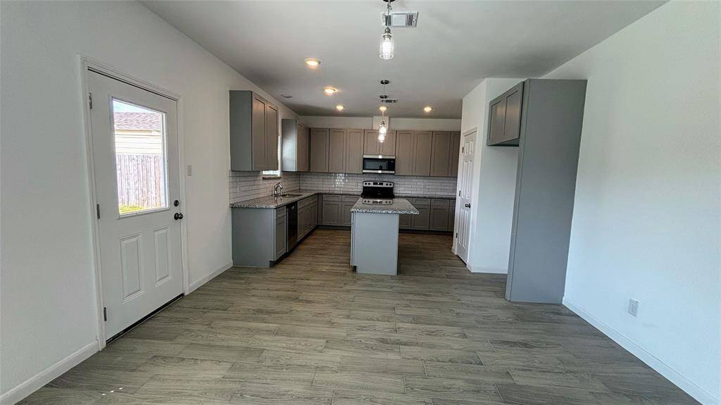The modern kitchen with sleek gray cabinetry, granite countertops, and stainless steel appliances. It features a central island, recessed lighting, and a light tile floor. A window leads to the outside, adding natural light to the space.