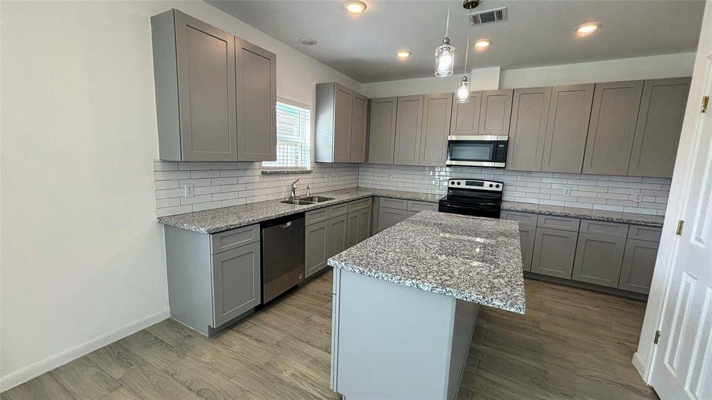 The modern kitchen with sleek gray cabinetry, granite countertops, and stainless steel appliances. It features a central island, recessed lighting, and a light tile floor. A window leads to the outside, adding natural light to the space.