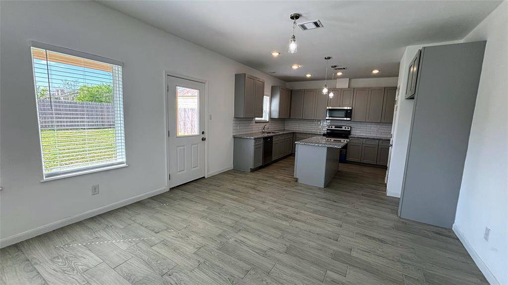 Bright, modern kitchen with gray cabinets, stainless steel appliances, and an island. Open layout with wood-style flooring and a view of the backyard through a large window.