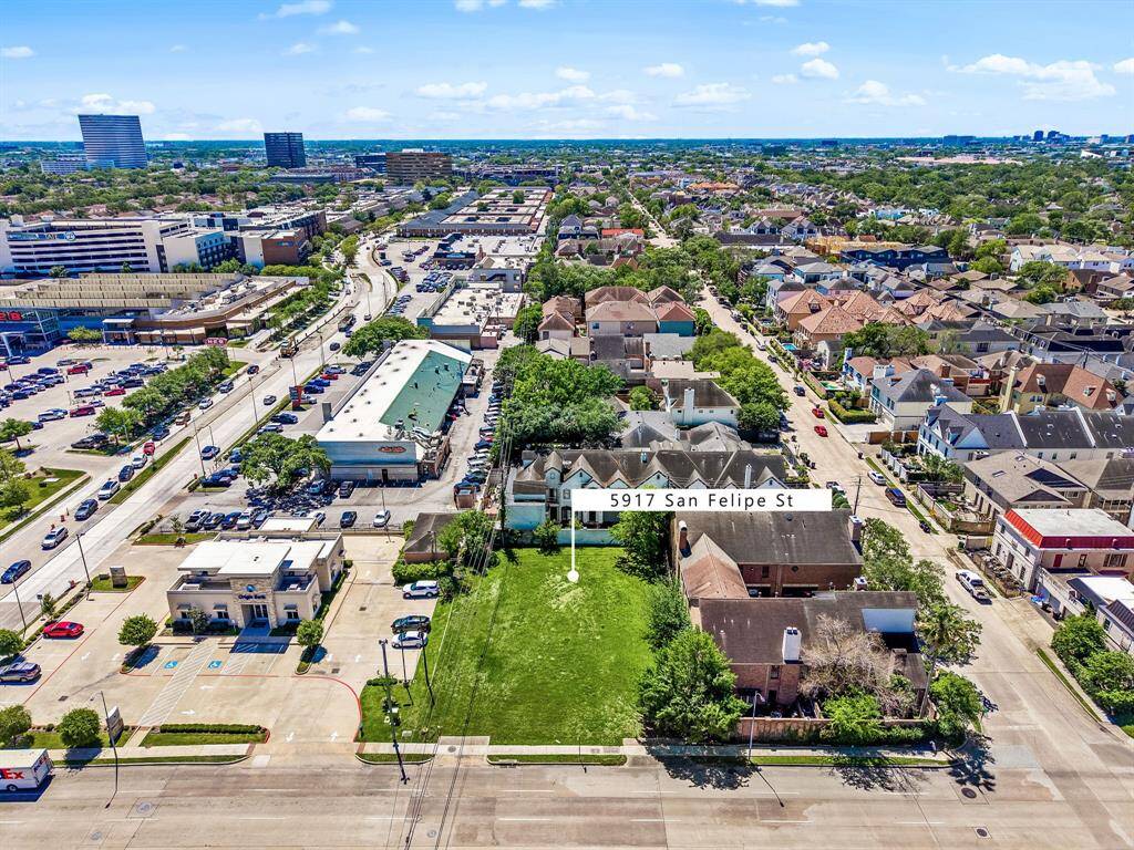 This aerial photo showcases a property at 5917 San Felipe St, highlighting a spacious green lot surrounded by a mix of residential and commercial buildings. The area appears bustling with nearby amenities, ample parking, and well-maintained homes, offering a vibrant urban environment.