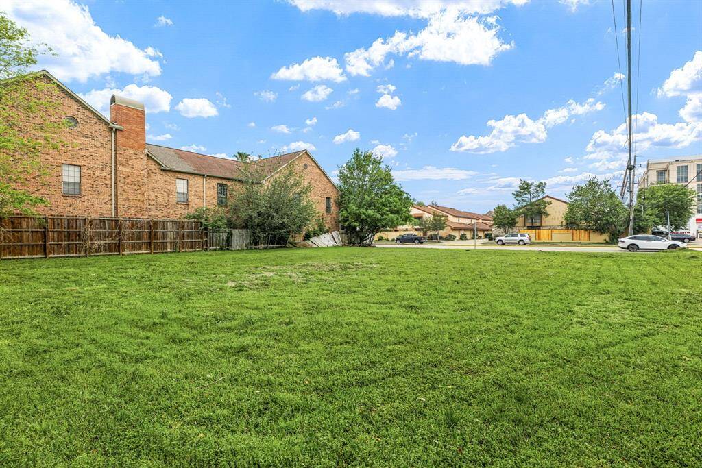 This photo shows a spacious, grassy lot with a backdrop of brick buildings and a fenced area. It's situated in a suburban neighborhood with nearby streets and parked cars, offering a mix of open space and urban convenience.