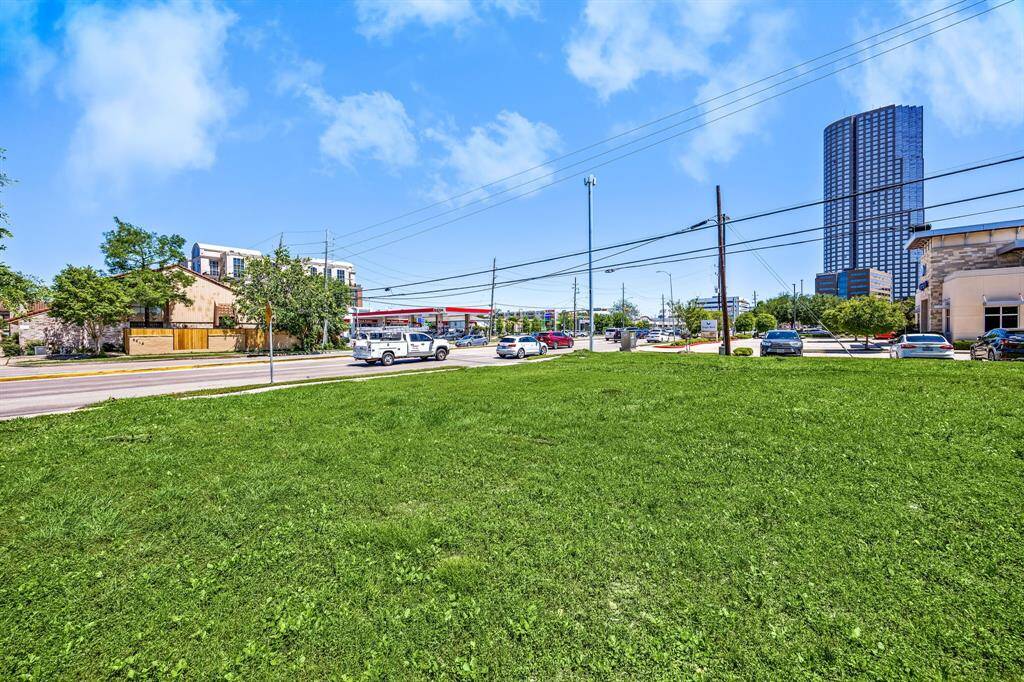 This photo features a spacious, grassy lot located near a busy street. The area is surrounded by residential and commercial buildings, with a mix of greenery, making it ideal for development or investment.