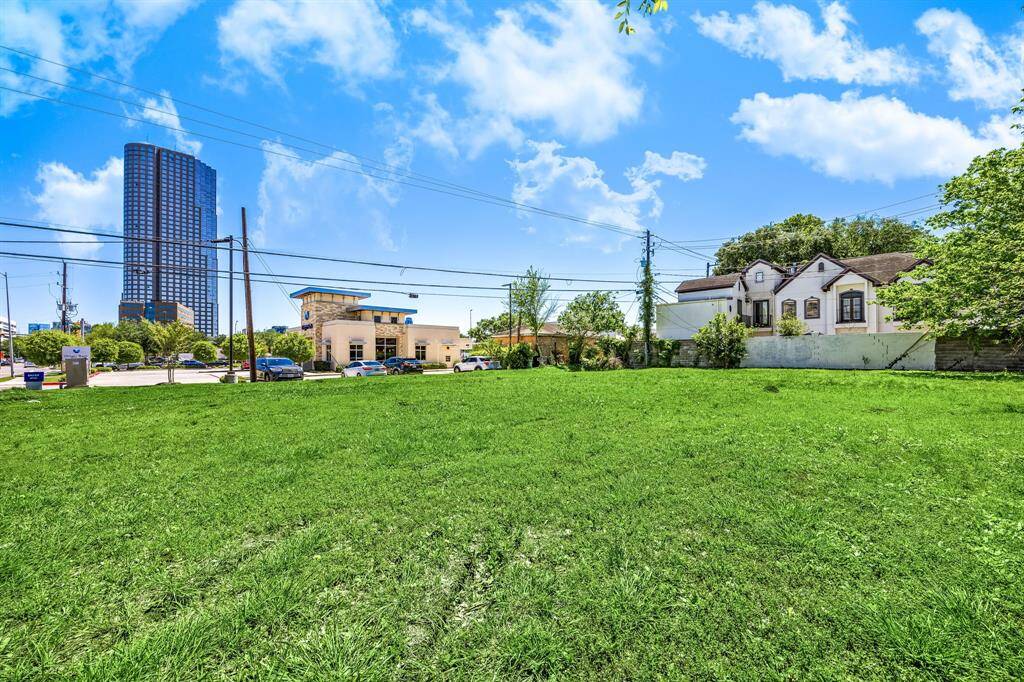 This photo features a spacious, grassy plot of land with a few nearby buildings, including a tall modern structure and a residential area. The setting is urban, with clear skies overhead, suggesting potential for development.