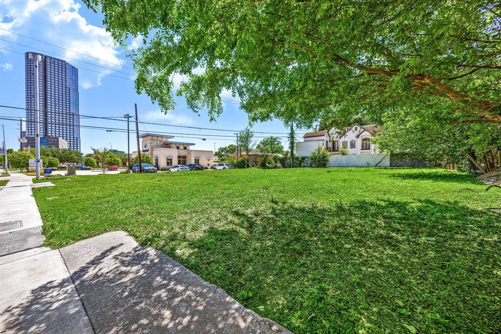 Spacious grassy lot with mature trees, adjacent to a sidewalk. Located near urban amenities, with a tall building and residential house visible nearby.