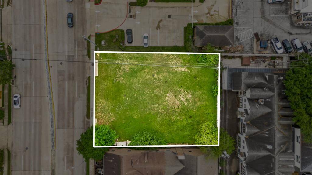 This aerial photo shows a rectangular plot of grassy land, bordered by roads and neighboring buildings. The area appears ready for development and is situated in a mixed-use neighborhood, offering convenient access to nearby amenities.