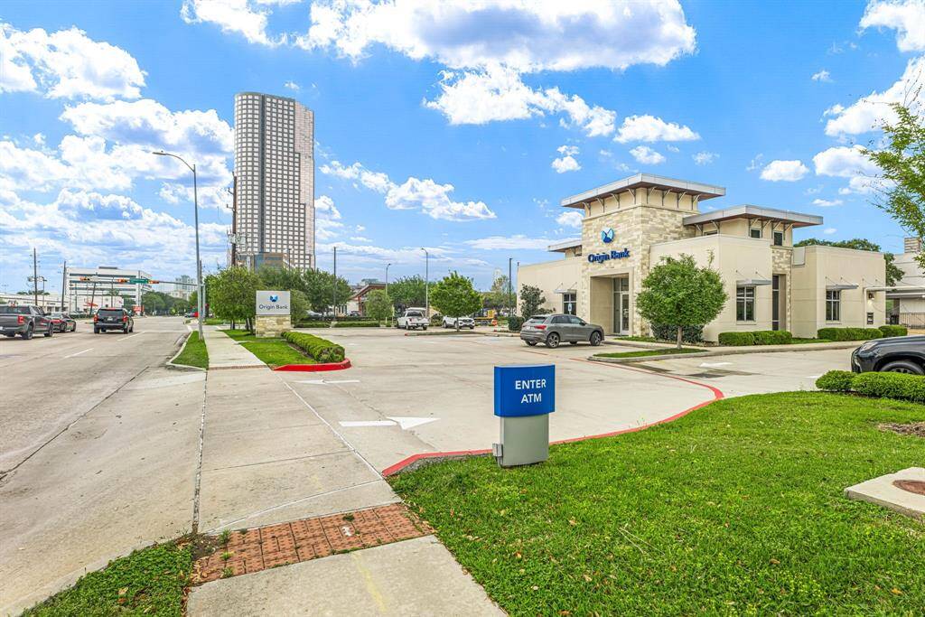 This photo shows a bustling urban area with a bank and ATM, located along a wide, well-maintained road. There's a mix of modern buildings, greenery, and ample parking, offering convenience and accessibility in a lively neighborhood setting.