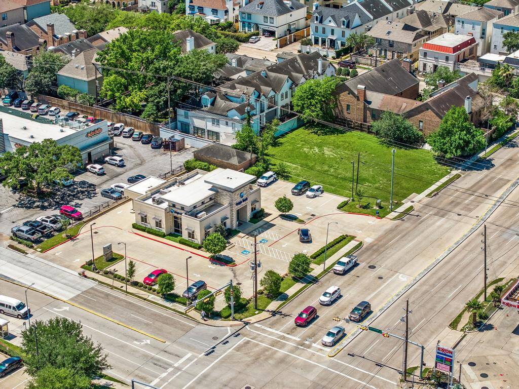 This aerial photo shows a suburban neighborhood with a mix of residential homes and commercial properties. In the foreground, there's a commercial building with parking, located on a busy intersection. Surrounding the area are well-maintained houses and green spaces, providing a blend of urban convenience and residential charm.