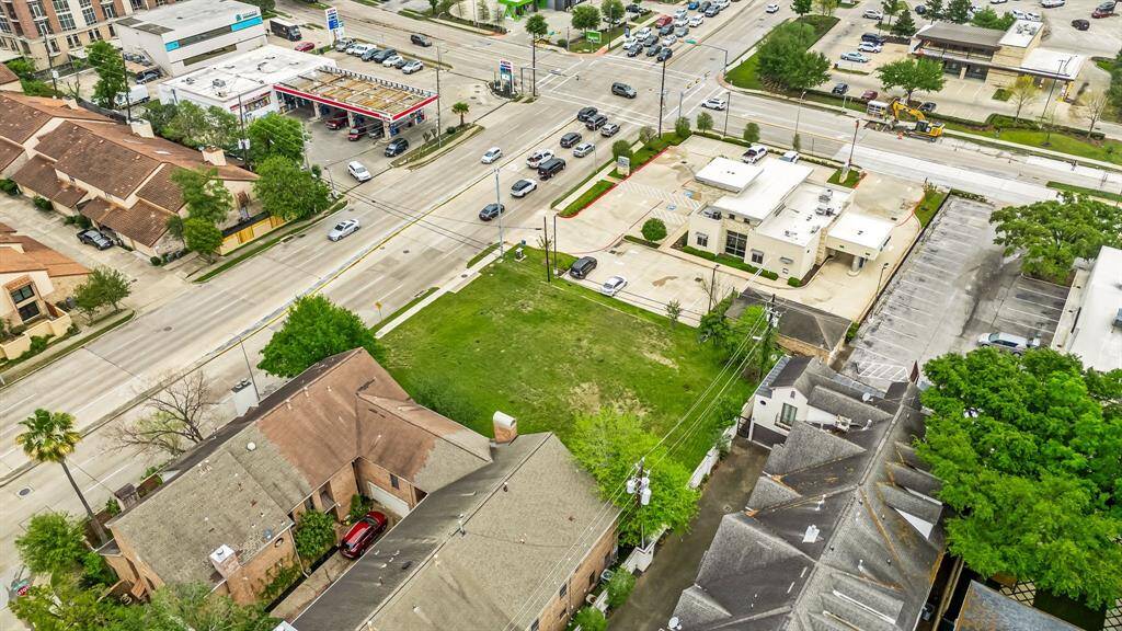 Aerial view of a commercial area with a vacant green lot, adjacent to residential buildings. Nearby are a gas station and several businesses, with a busy intersection. Ideal for potential development or investment.