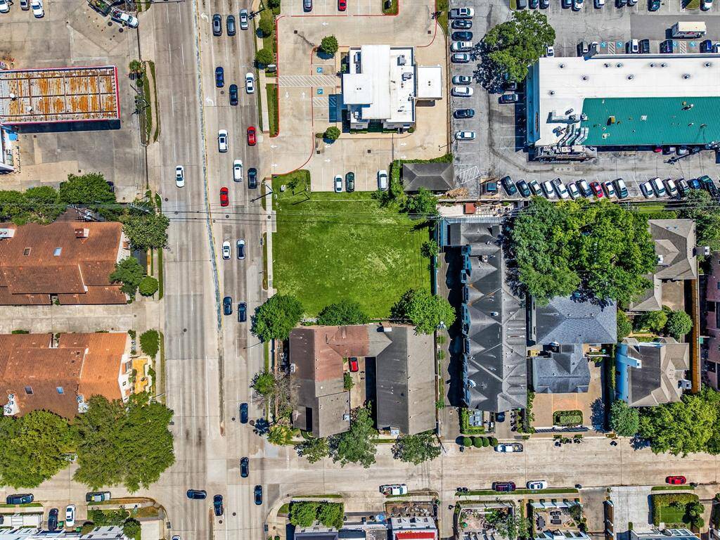 This aerial photo showcases a neighborhood with a mix of residential and commercial buildings. There's a large green plot in the center, surrounded by roads with moderate traffic and ample parking. The area appears well-maintained, offering convenient access to nearby businesses and amenities.