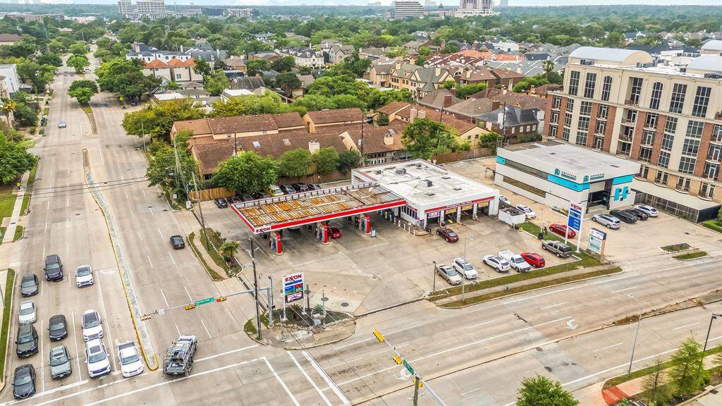 This aerial photo shows a neighborhood with a mix of residential and commercial properties. It features a gas station on a busy street corner, surrounded by houses and a few larger buildings. The area appears to be well-developed with tree-lined streets and ample parking.