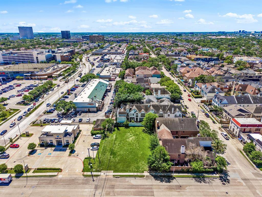 This photo shows a vacant lot in an urban neighborhood, bordered by residential buildings and commercial establishments. It offers a clear, grassy space ideal for new construction, with convenient road access and nearby amenities.