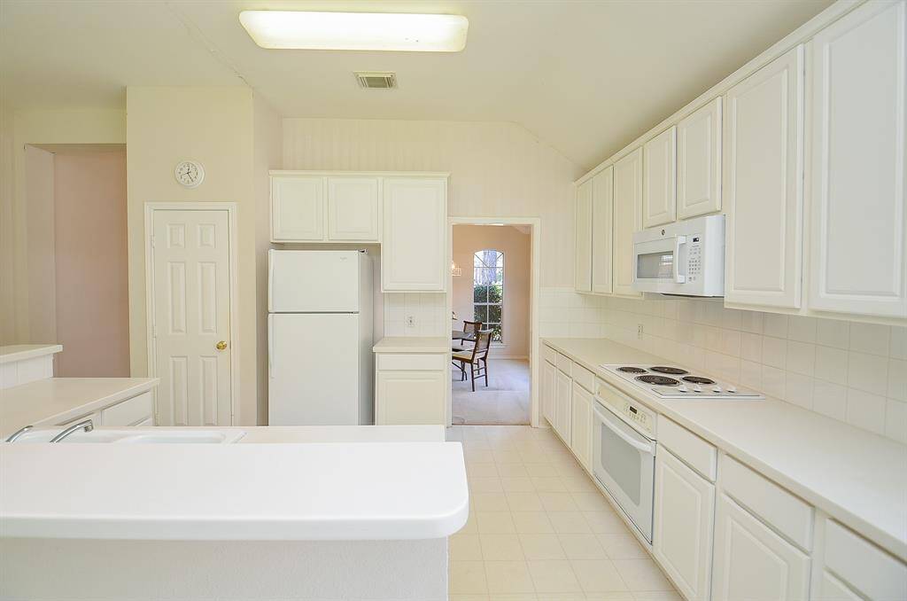 Bright kitchen with white cabinets, countertops, and appliances, including a built-in stove and microwave. The space is open with a view into the dining area, featuring a large window for natural light.