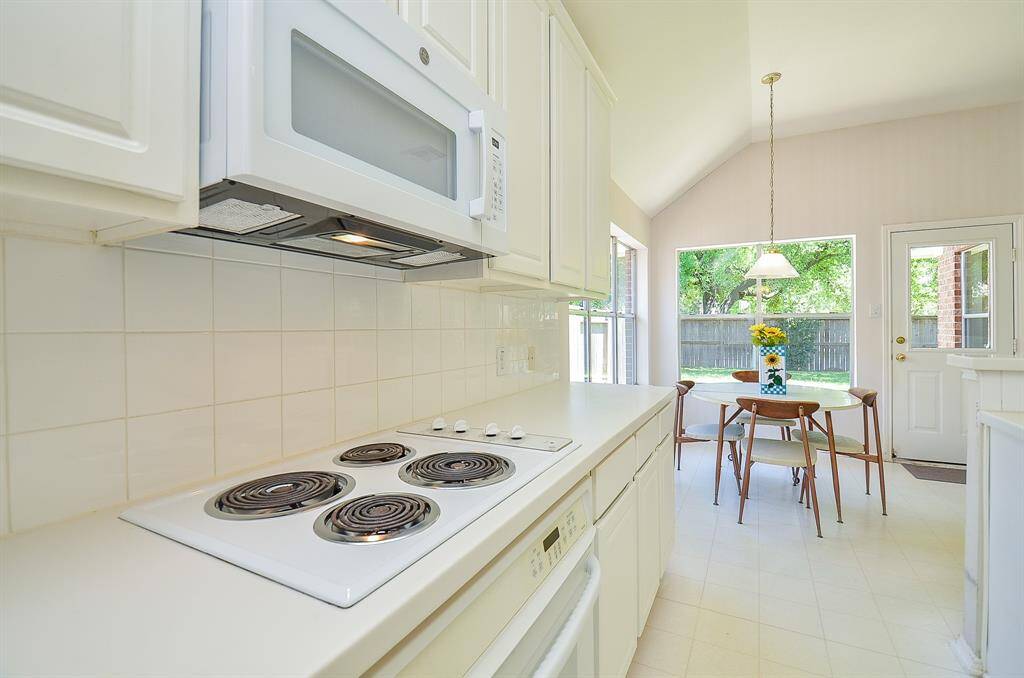 Bright kitchen with white cabinetry, tile backsplash, and electric stovetop. Adjacent dining area features large windows and a door leading to the backyard, allowing plenty of natural light.