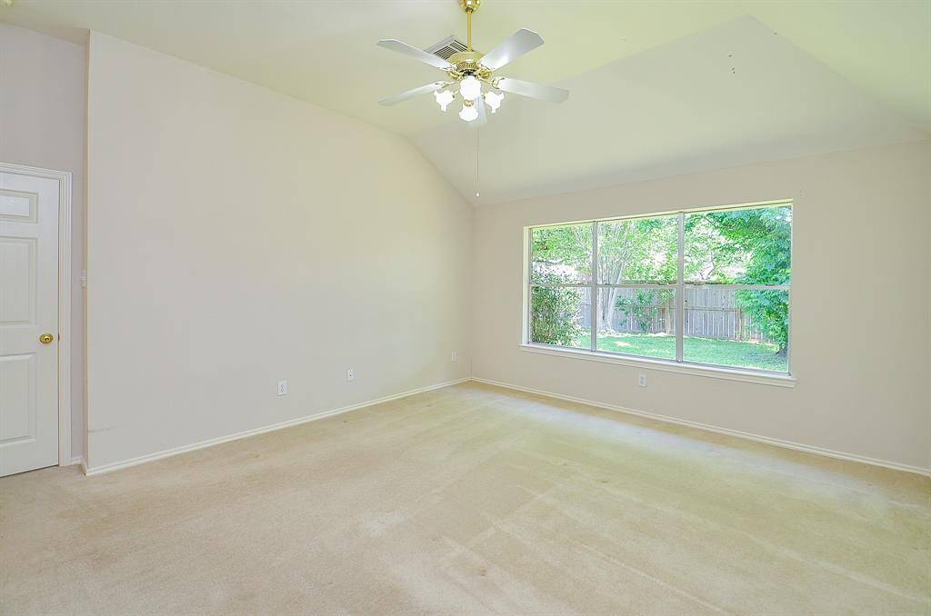 This spacious, primary bedroom room features a large window offering plenty of natural light and a view of the green backyard. It has a ceiling fan with light fixtures and neutral walls, making it versatile for any decor style.