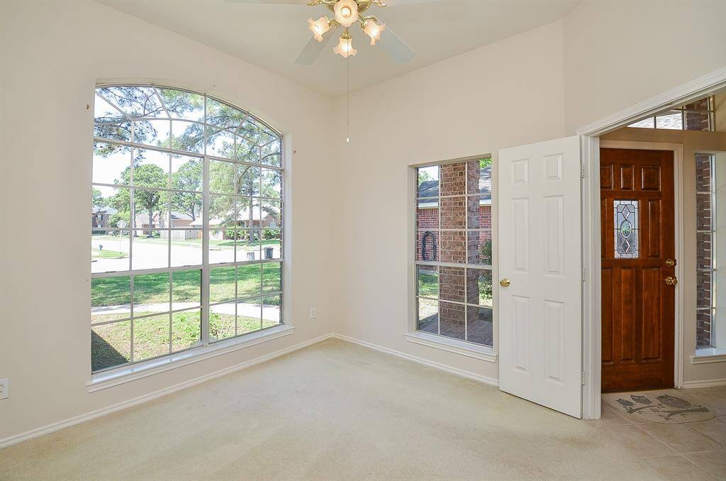 This bright room features large windows with a picturesque view of the neighborhood, a ceiling fan with lights, and neutral walls. The adjacent doorway with a glass-accented wooden door enhances the space's welcoming feel. Use as a home office, or 4th bedroom if needed!