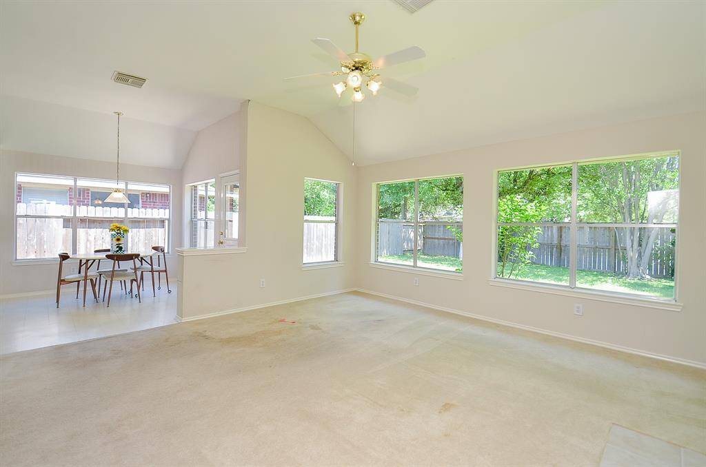 This bright, open living space features large windows overlooking a fenced backyard, with a dining area adjacent. The room has high ceilings, a ceiling fan, and a neutral color palette, offering a versatile layout for various decor styles.