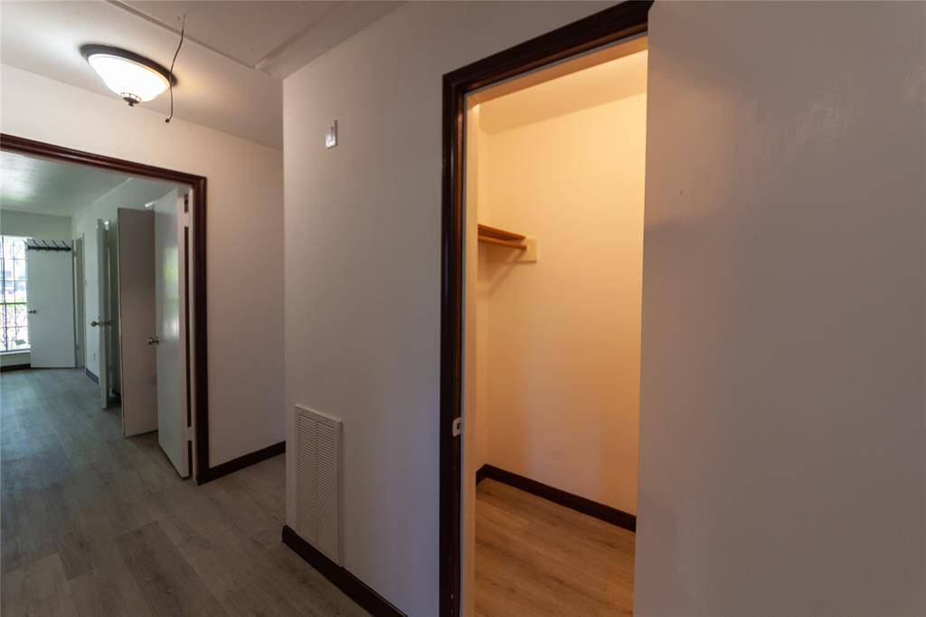 A hallway with laminate flooring, leading to a well-lit room with a window. Adjacent to the hallway is a spacious closet with wooden shelving. The space is clean and has a modern light fixture.