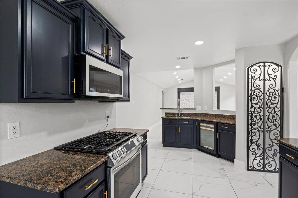 This kitchen features dark cabinetry with brass handles, a modern stainless steel oven and microwave, and a granite countertop. The space includes white tile flooring and a decorative wrought iron gate, adding a touch of elegance.