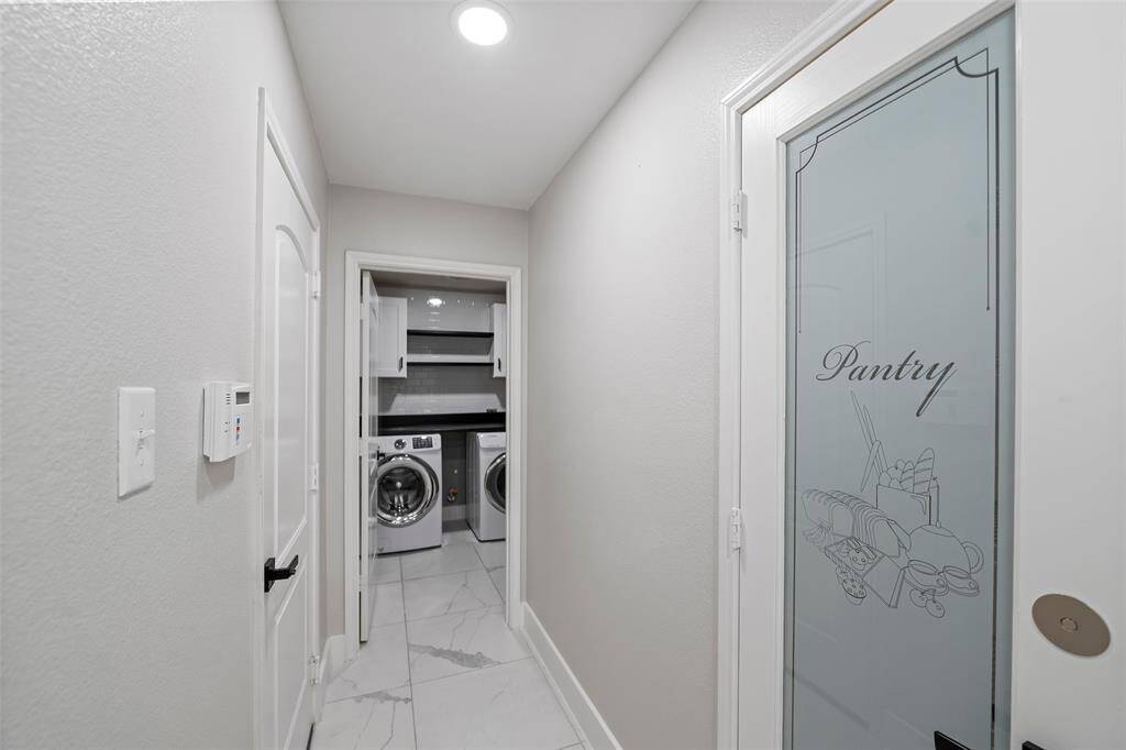 The photo shows a bright, modern hallway with a frosted glass pantry door on the right and a laundry area at the end. The laundry has a washer and dryer with shelving above, and the hallway features sleek tile flooring and recessed lighting.