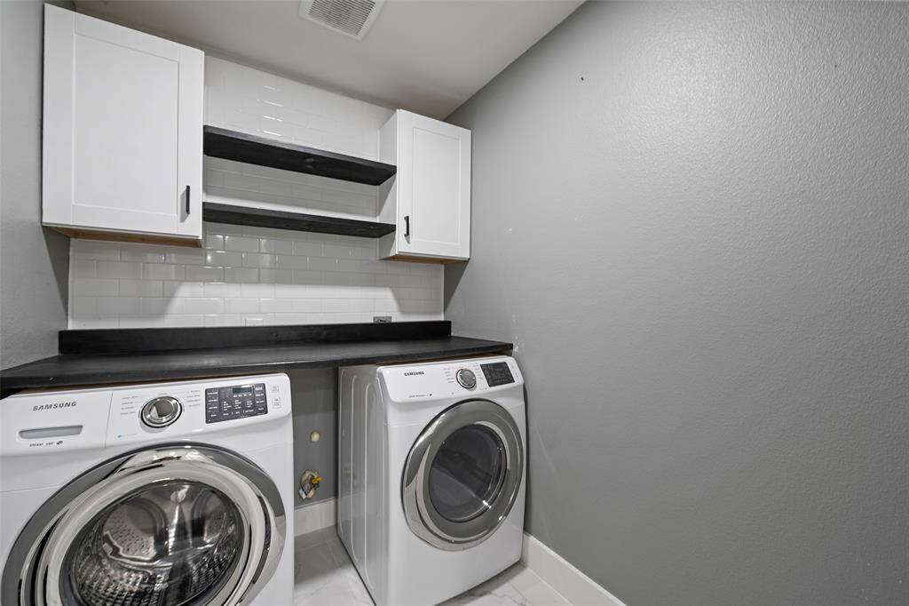 This photo shows a modern laundry room with a Samsung washer and dryer, sleek white cabinetry, and open shelves. The walls are painted gray, complemented by a white subway tile backsplash.