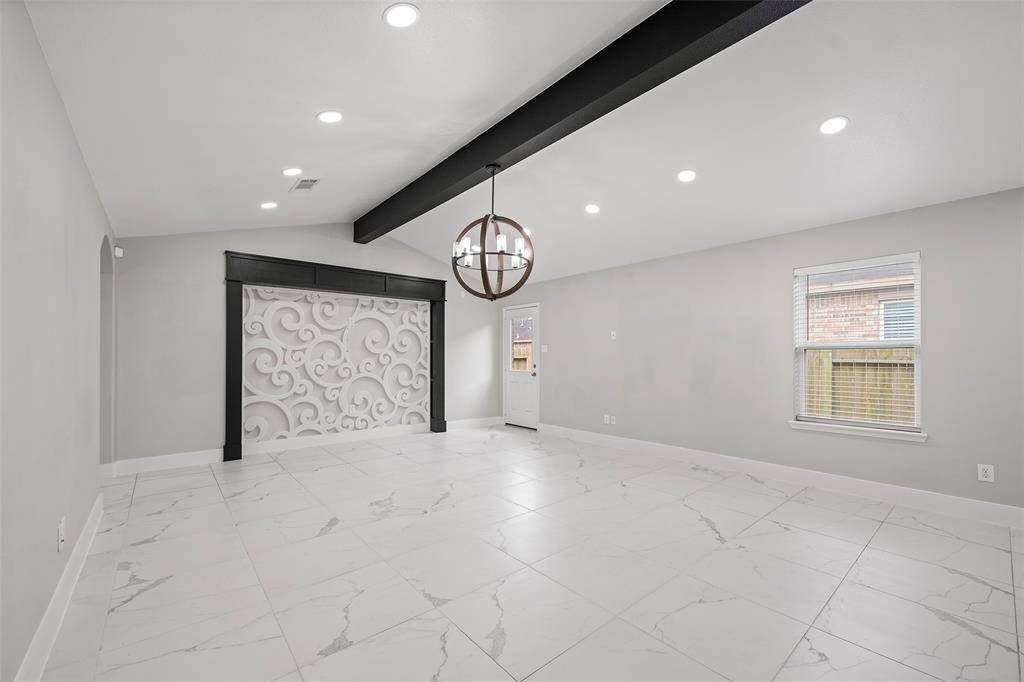 This spacious room features a modern design with marble-patterned tile flooring, a decorative wall panel, and a striking black ceiling beam. It includes recessed lighting and a large window for natural light. A contemporary chandelier adds elegance to the space.