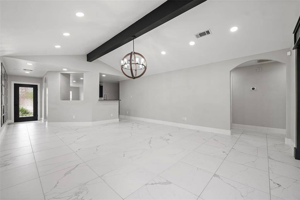 This spacious, modern living area features sleek white tile flooring, recessed lighting, and a stylish chandelier. An open layout connects to the kitchen, and large windows bring in natural light, enhancing the airy atmosphere.