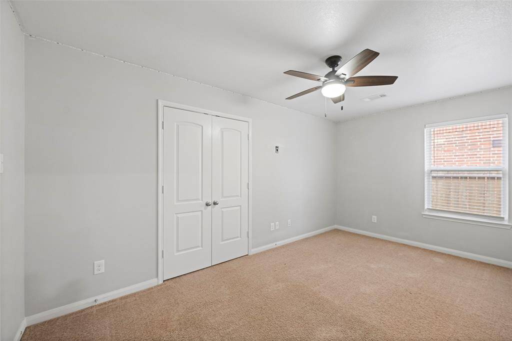 This is a cozy bedroom with neutral-toned walls and carpet, featuring a ceiling fan with lights. It has a window for natural light and a double-door closet for storage.