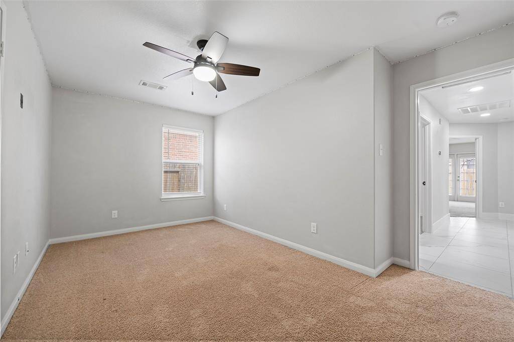 This room features soft carpeting, a modern ceiling fan, and neutral walls. It includes a window for natural light and leads to a tiled hallway. Perfect for a cozy bedroom or office space.