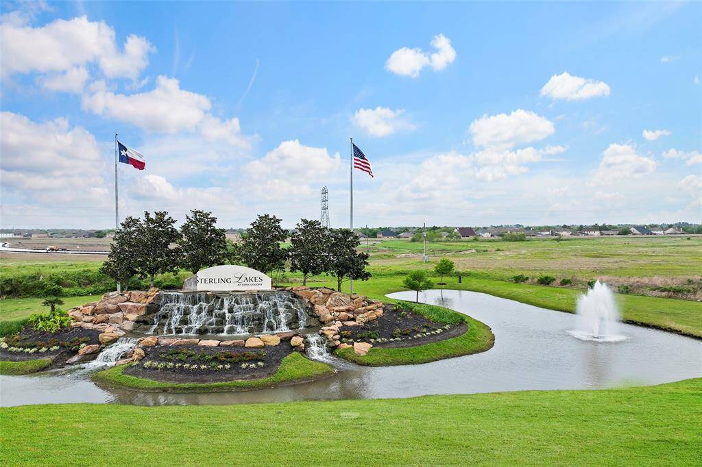 The photo showcases a picturesque entrance to a residential community, featuring a cascading waterfall surrounded by manicured landscaping. There's a pond with a fountain, and flags are prominently displayed, creating a welcoming and serene atmosphere.