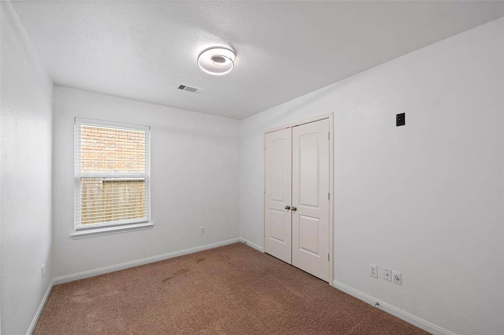 This photo shows a small, carpeted bedroom with white walls. It features a modern ceiling light, a window with blinds, and a double-door closet, offering a cozy and functional space.