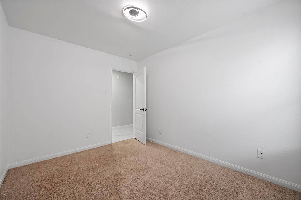 This photo showcases a small, carpeted room with white walls, a modern ceiling light, and an open door leading to a tiled area, providing a clean and versatile space.