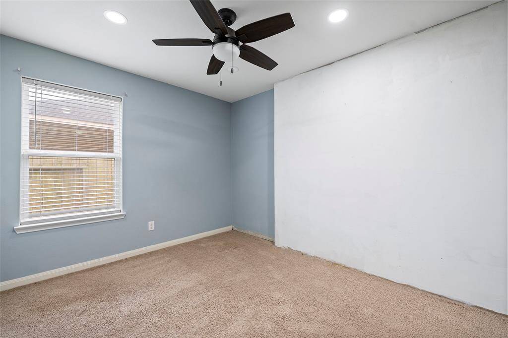 This room features soft blue walls and a beige carpet, complemented by a ceiling fan and recessed lighting. A window with blinds allows natural light to fill the space, offering a cozy and functional environment.