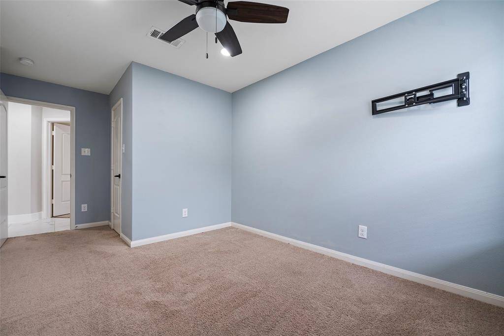 This room features soft blue walls, beige carpet, and a ceiling fan. It has a wall mount for a TV and a view into an adjacent hallway with white doors.