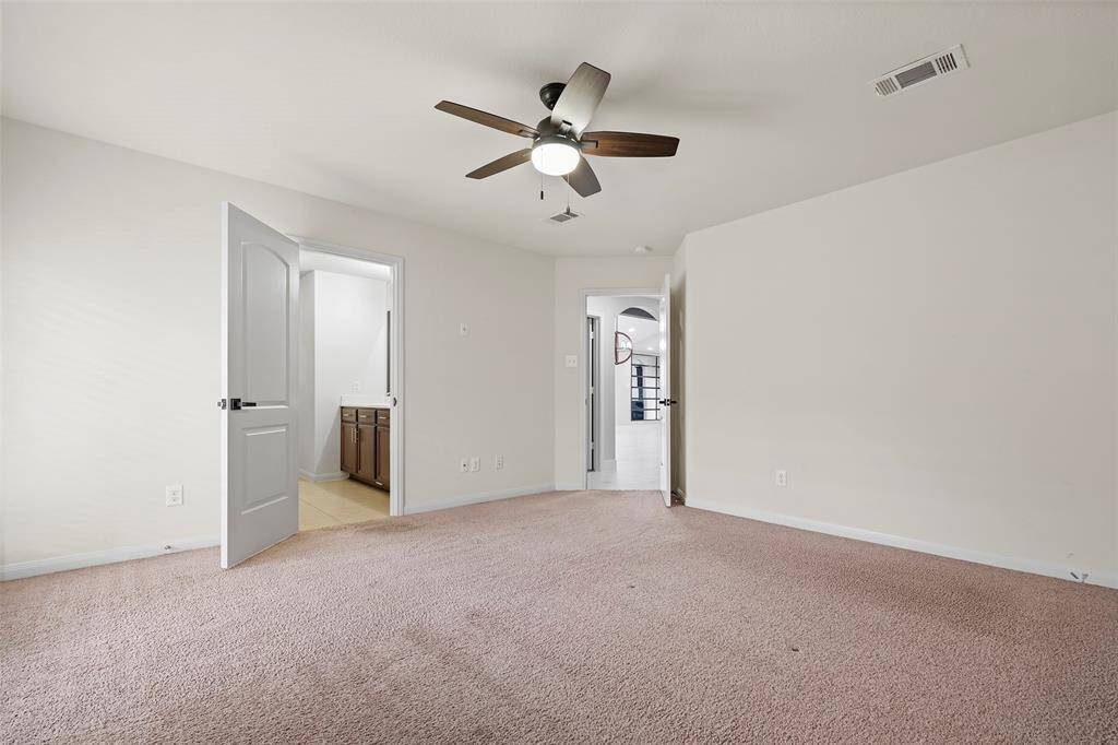This spacious bedroom features neutral carpeting, a ceiling fan, and ample natural light. It connects to a bathroom with visible cabinetry and has a second doorway leading to another part of the house.
