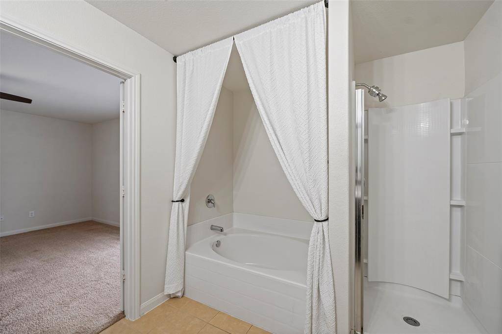 The photo shows a bathroom with a white tub and shower combination. The tub is partially enclosed by a white curtain. The bathroom connects to a carpeted room, likely a bedroom, through an open doorway. The space is clean and has a neutral color palette.