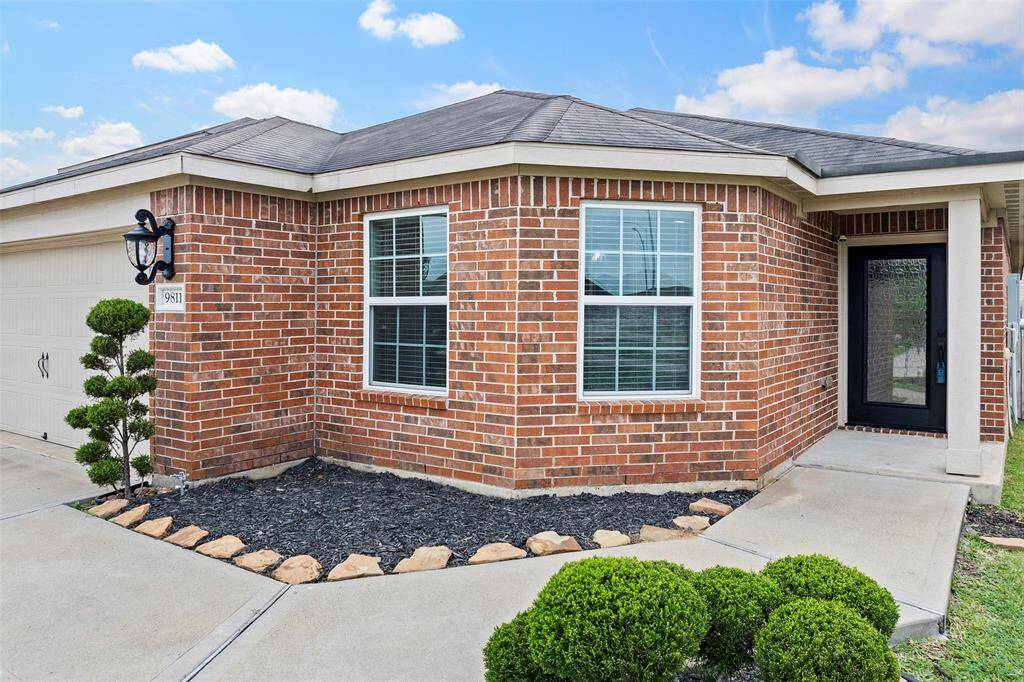 This photo showcases a charming single-story brick home with a modern entryway. It features a well-maintained front yard with decorative shrubs and a paved walkway leading to the front door. The exterior is complemented by a two-car garage and a stylish black front door.