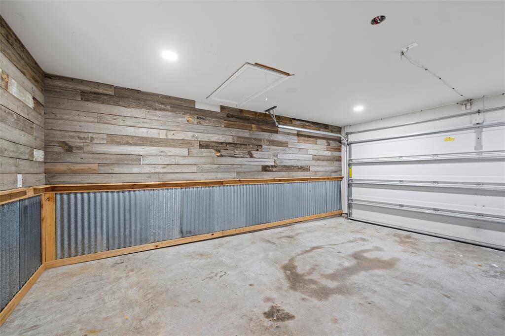 This garage features rustic wood paneling and corrugated metal accents, providing a cozy, industrial feel. It has a concrete floor, overhead lighting, and a large garage door, offering ample space for parking or storage.