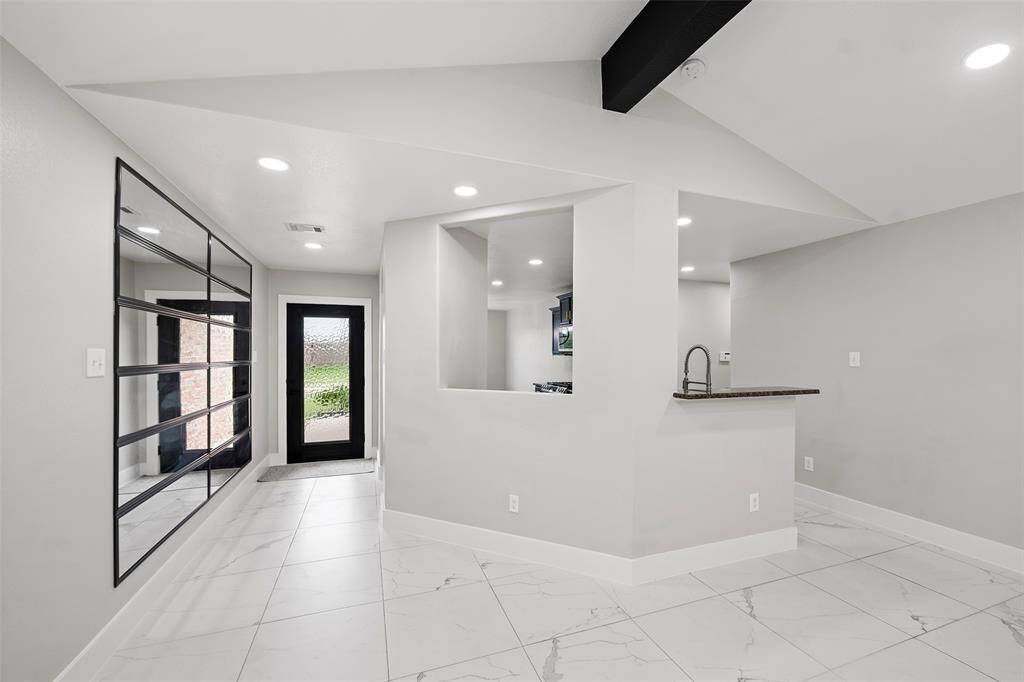 This modern hallway features sleek tile flooring, recessed lighting, and a wall of mirrors. It leads to a kitchen with a stylish pass-through window and a black-framed glass door, offering a bright and open feel.