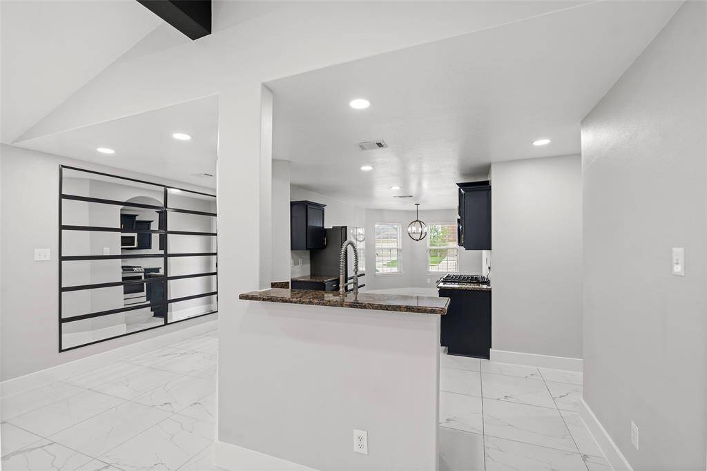 This modern kitchen features sleek black cabinetry, a granite countertop, and stainless steel appliances. It opens to a bright dining area with large windows, and the space is accented by elegant tile flooring and recessed lighting. A unique partition with horizontal slats adds a stylish touch.