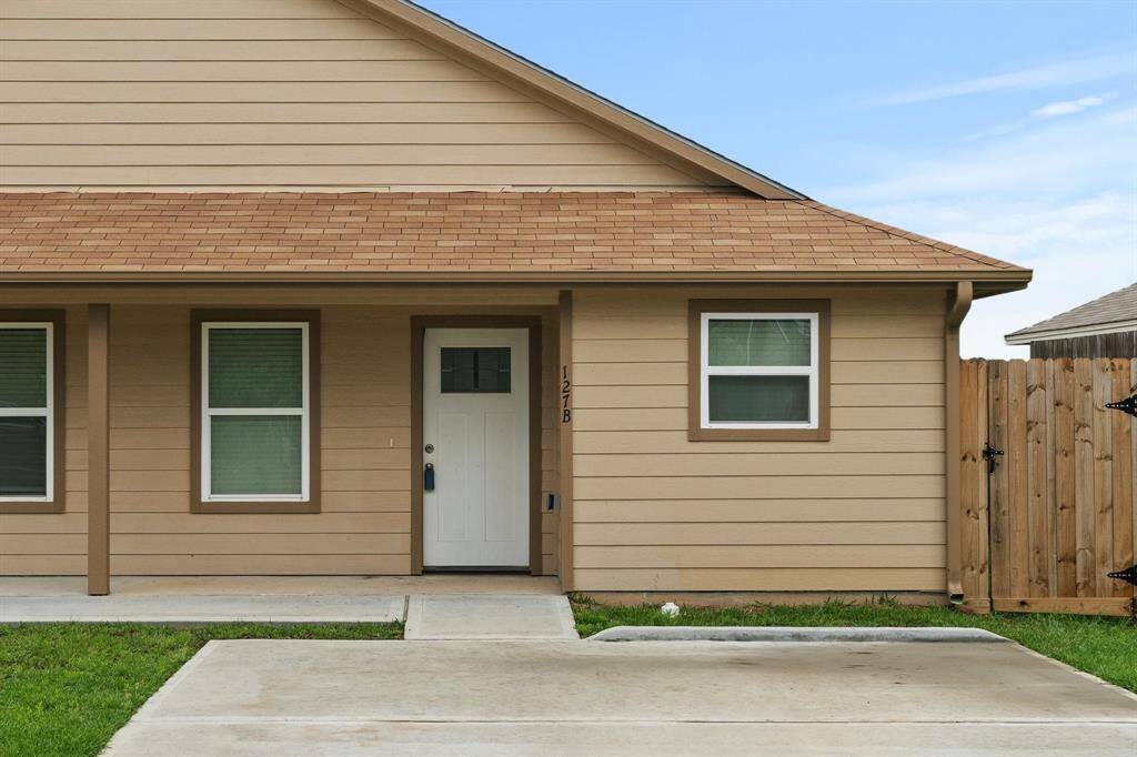 This is a single-story home featuring a tan exterior with white trim, a shingled roof, and a concrete walkway leading to the front door. There are two front windows with blinds, and a privacy fence. Yard Maintenance included in your lease.