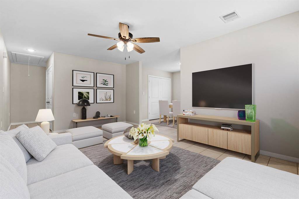 This is a modern, well-lit living space featuring a comfortable seating area, a ceiling fan, and a large flat-screen TV. The room has a neutral color scheme and includes a small work area. The open layout extends towards a dining area visible in the background.