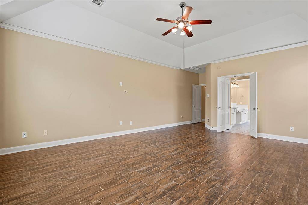 This spacious primary bedroom room features warm-toned tile flooring, neutral walls, and a ceiling fan. It has ample natural light and leads into a bathroom through double doors, providing a bright and airy feel.