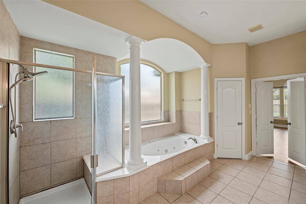 This spacious bathroom features a large corner shower with frosted glass, a luxurious soaking tub with elegant columns, and neutral tile flooring. Double doors lead to the adjoining primary bedroom, enhancing the open layout.