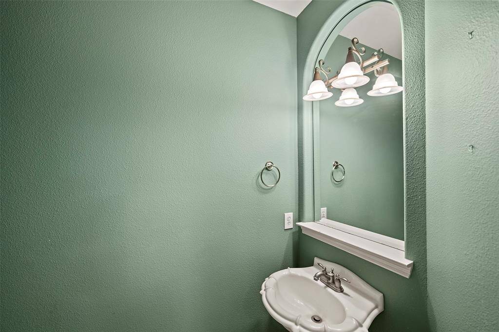 A half bathroom with pastel green walls, featuring a decorative pedestal sink and an arched mirror with an elegant light fixture above.